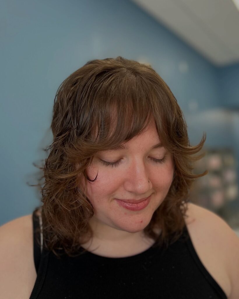 A woman with a caramel brown wolf cut hairstyle featuring soft waves and curtain bangs.