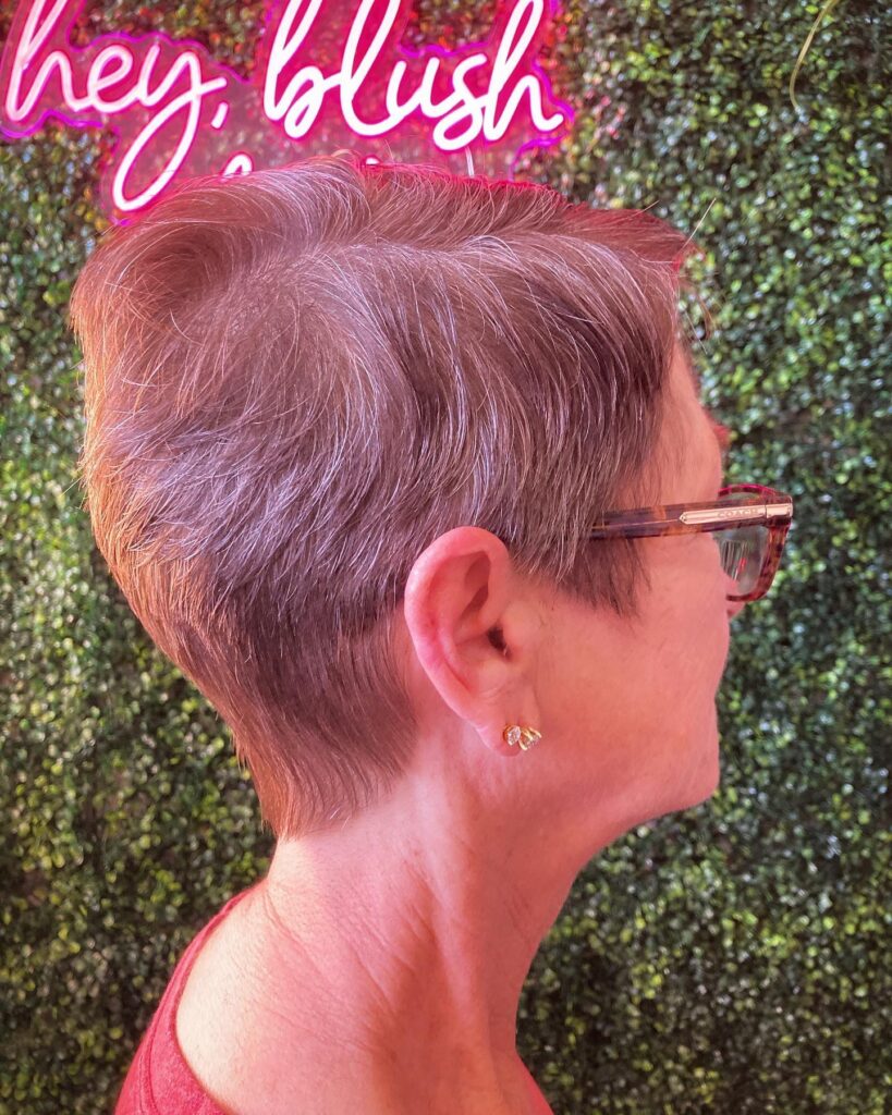 A woman with short, naturally gray hair in a refined, sleek, tapered cut, viewed from the side, wearing tortoiseshell glasses.