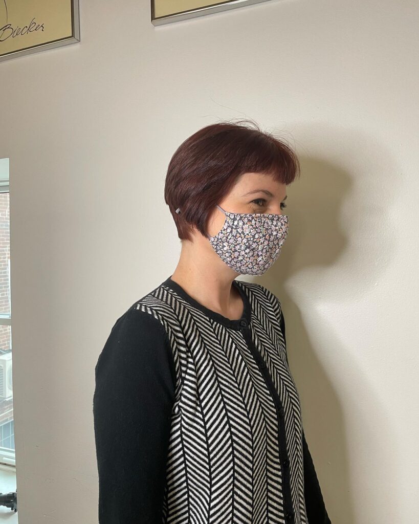 A woman with a rich auburn short bob haircut featuring a distinct side taper and subtle micro bangs, viewed from a side profile while wearing a patterned face mask.