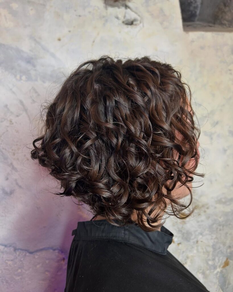 A side profile of a woman with a glossy dark brown curly bob featuring a tapered back and bouncy, defined ringlets.