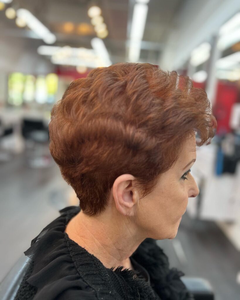 A side profile of a woman with a wavy copper pixie cut featuring a neat tapered nape and volume on top.
