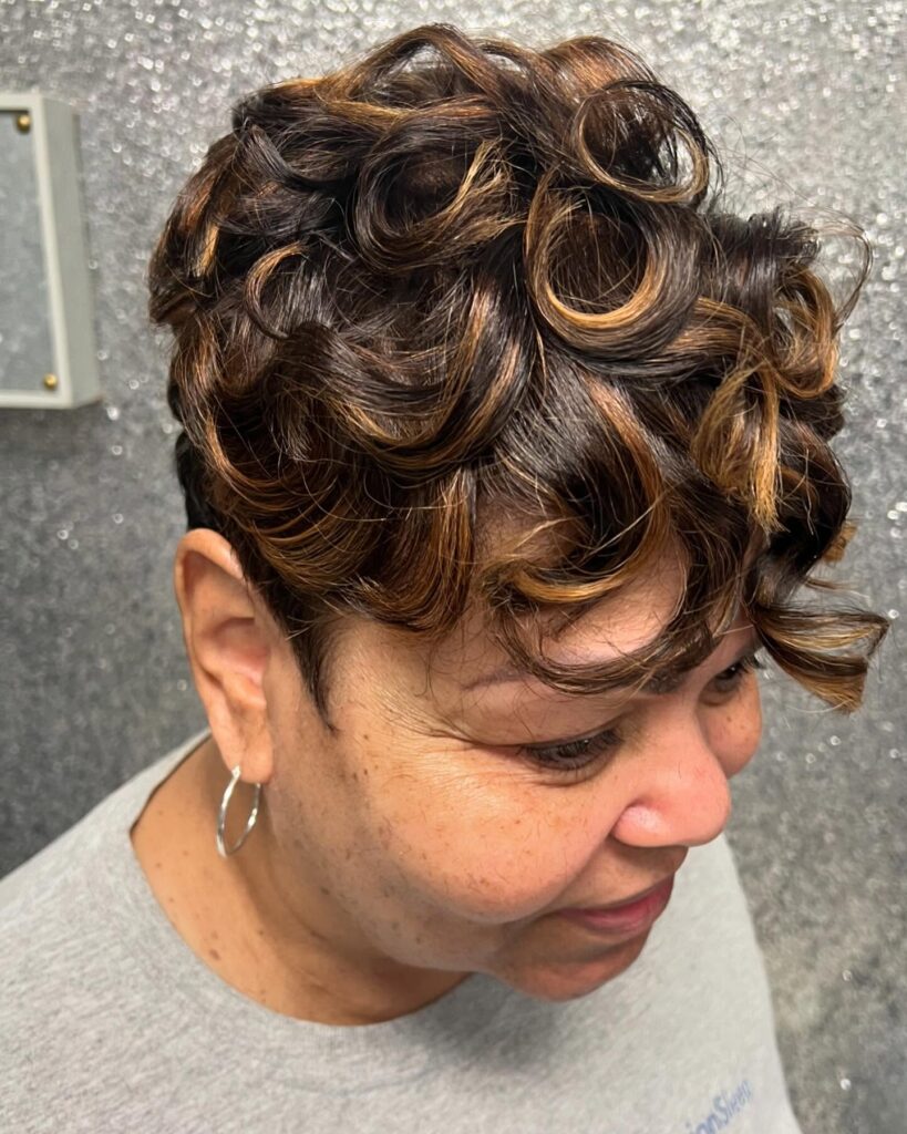 A woman with short, voluminous curls featuring caramel highlights and a sleek tapered side and back.