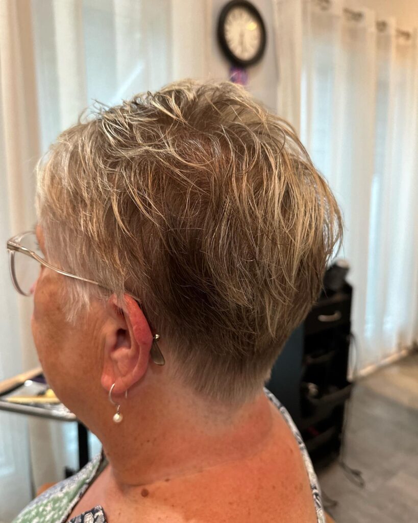 A sideback view of a woman with a wispy ash blonde pixie cut featuring feathered layers and a very short, clean tapered nape.