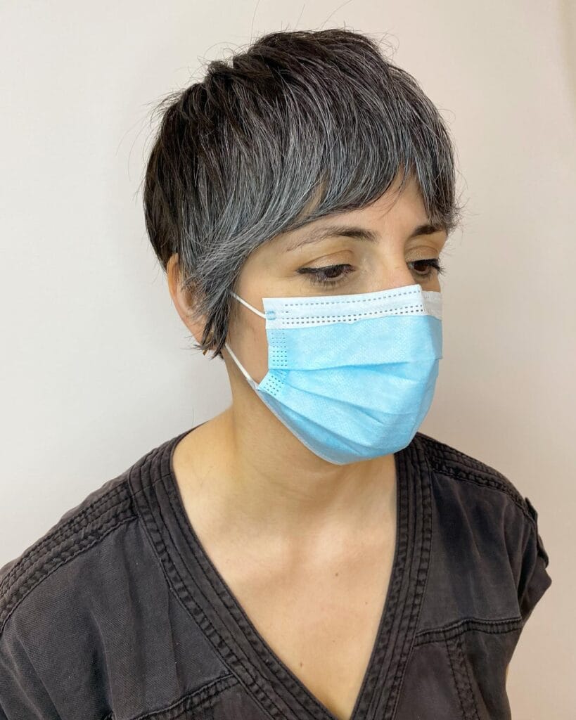 A woman with a short, textured, salt-and-pepper pixie cut and side-swept bangs wearing a blue face mask.