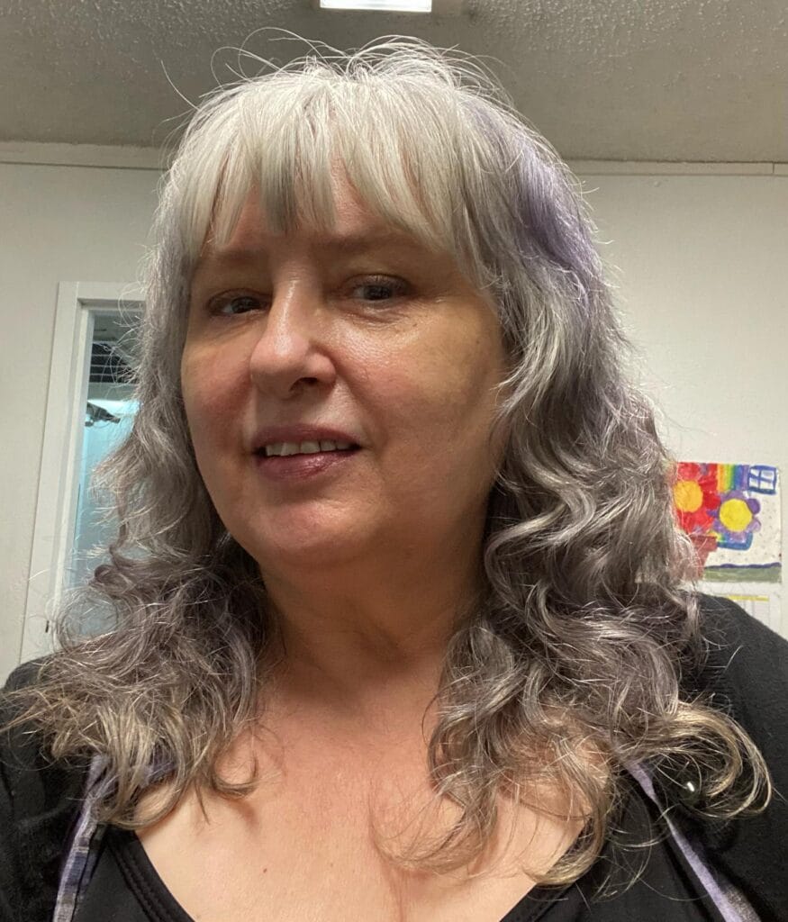A woman with long, curly silver and lavender hair and a full, straight fringe is smiling for a selfie.