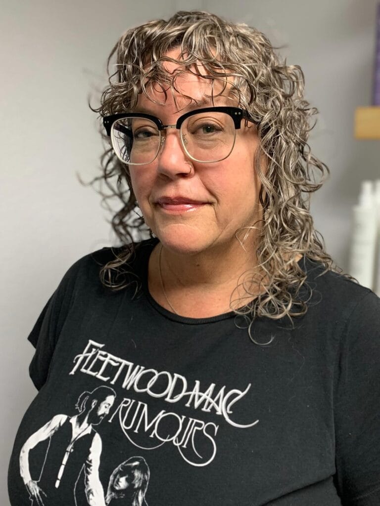 A woman with shoulder-length curly brown hair and soft piecey bangs wearing black glasses and a graphic t-shirt.
