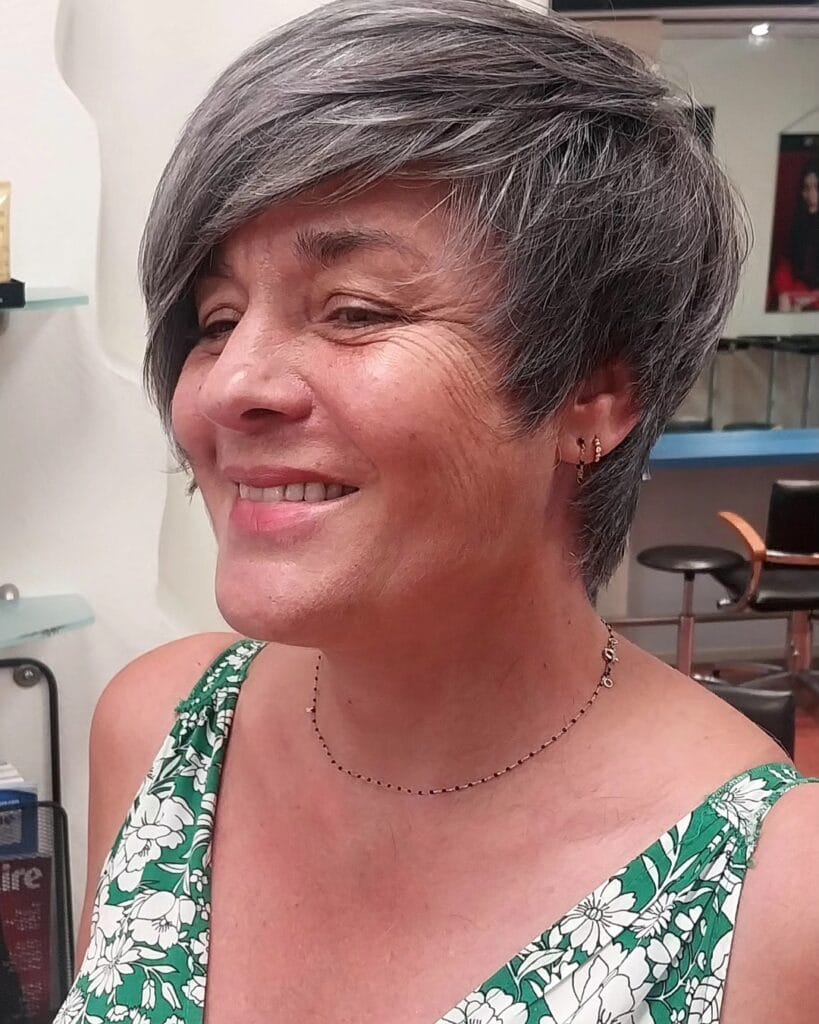 A smiling woman with a textured charcoal and silver pixie cut featuring a long side-swept fringe.