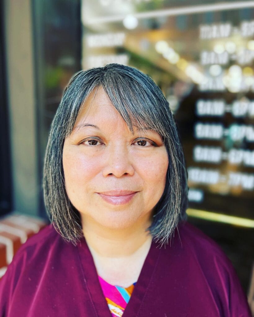 A woman with a straight, salt-and-pepper bob and side-swept bangs is looking forward and smiling.