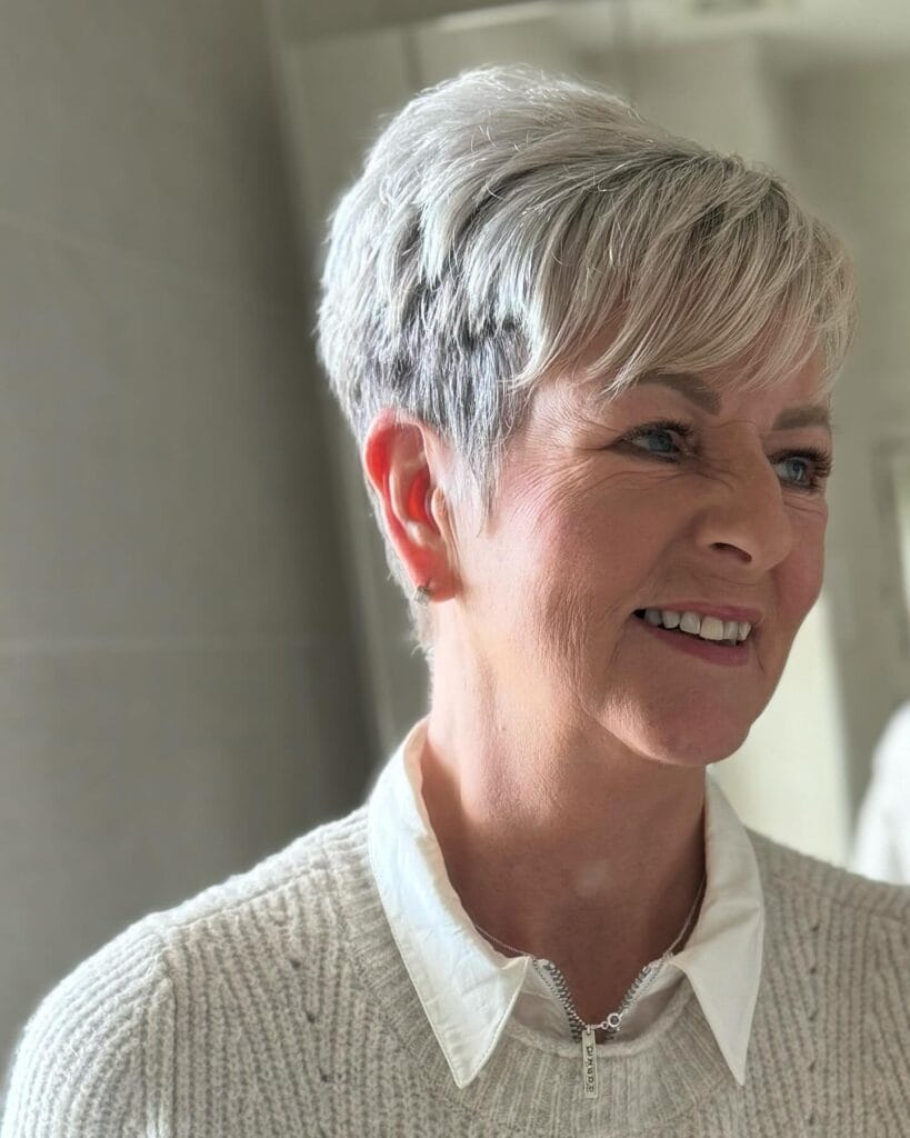 A smiling woman with a short textured silver pixie cut and soft side-swept bangs is looking to the side.