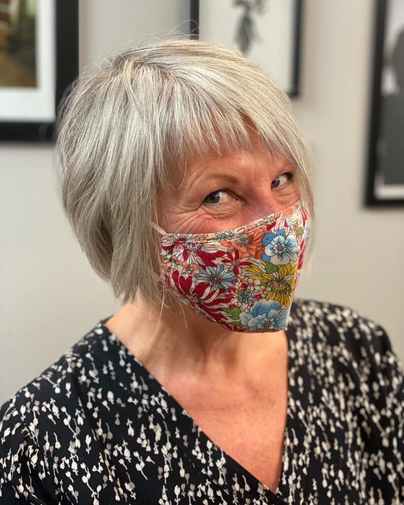 A woman with a soft, feathered silver bob and short piecey bangs wearing a floral face mask.