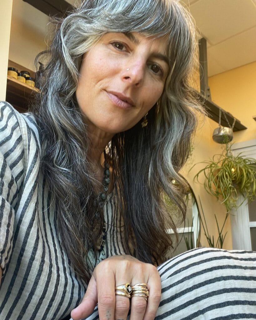 A woman with long layered salt-and-pepper hair and thick feathered bangs looking at the camera.