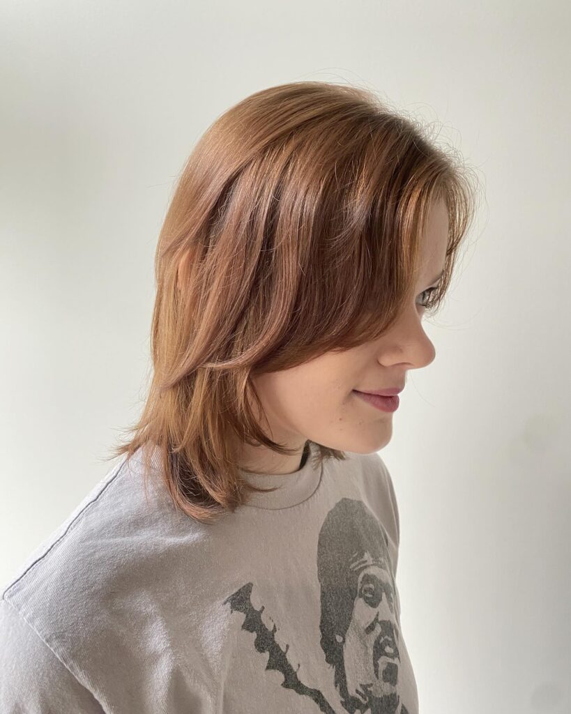 A profile view of a woman with warm caramel brown hair featuring long, sweeping layers and a side-swept fringe.