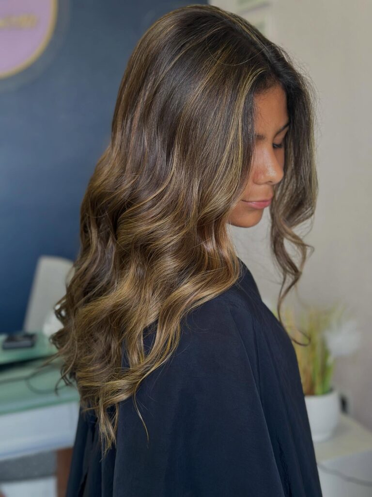 A woman with medium length dark brown hair featuring golden blonde highlights styled in soft voluminous waves.