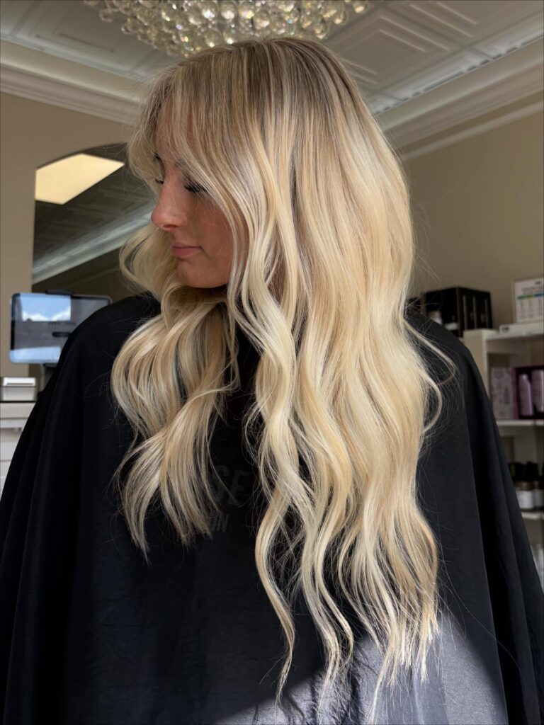 A woman with long buttery platinum blonde hair styled in soft waves with wispy face framing bangs.