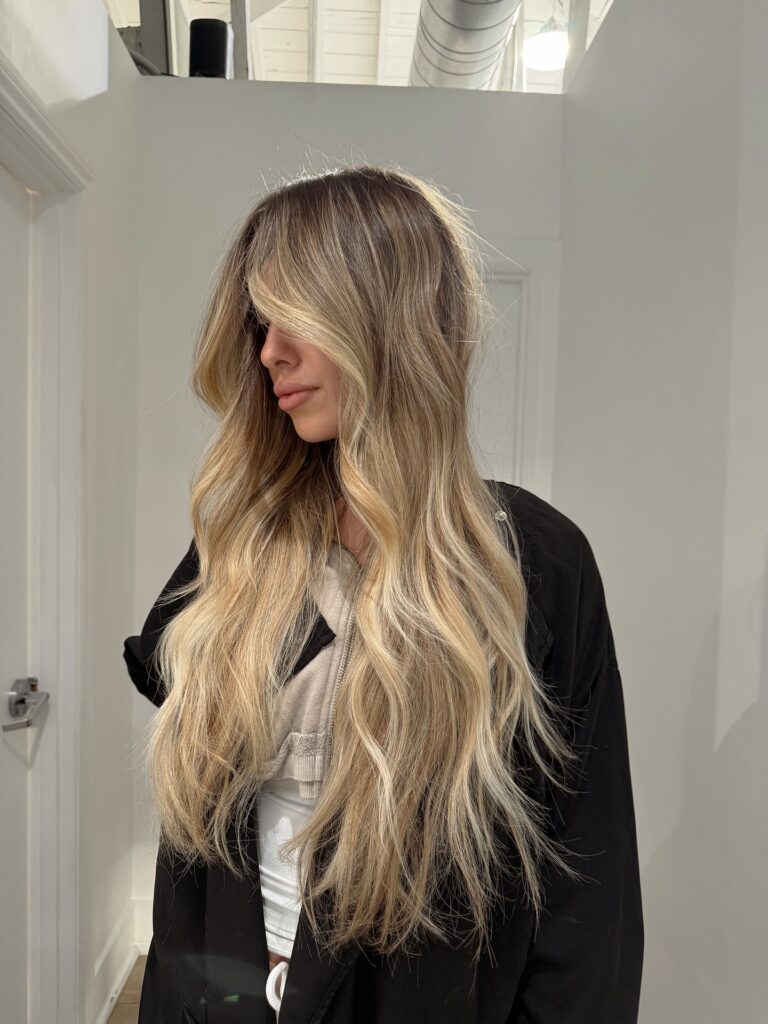 A woman showing off her long honey blonde balayage hair with wavy texture and face framing layers.