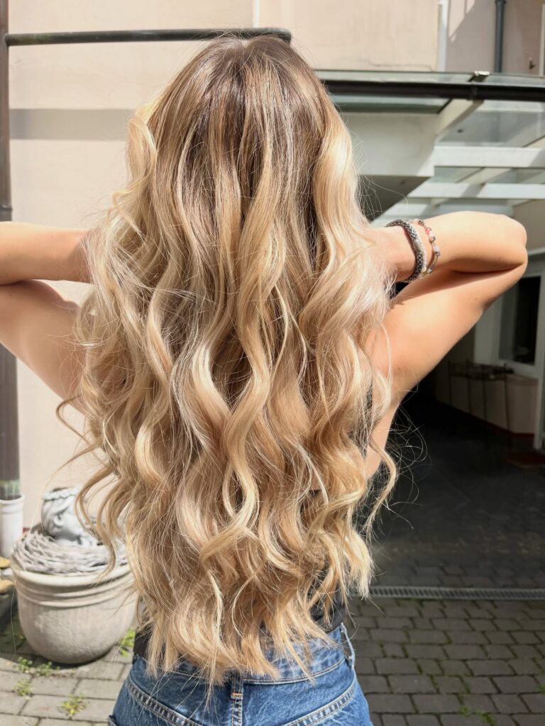  Rear view of a woman with long sandy blonde hair featuring dimensional balayage styled in soft flowing waves.