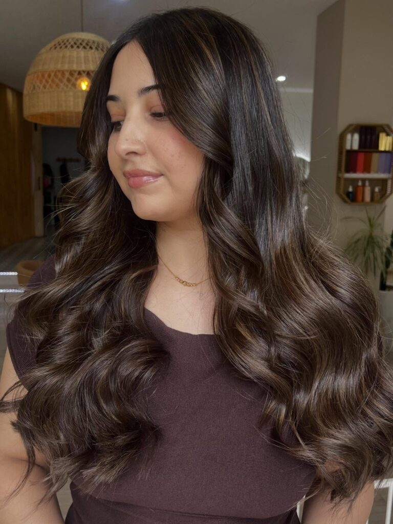 A woman with long dark espresso brown hair styled in voluminous glossy waves with soft face framing layers.