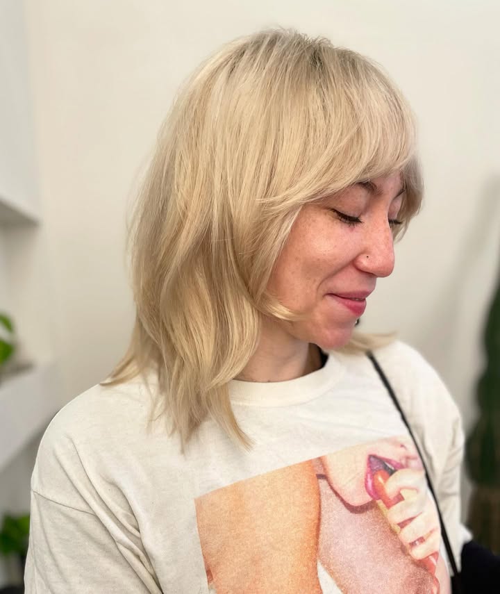 Chin length blonde bob with textured ends and full bangs across the forehead.