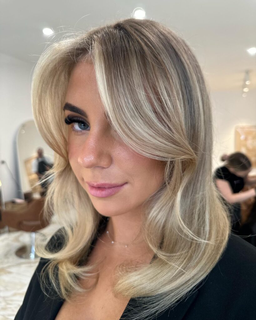 A woman with a shoulder length blonde butterfly haircut featuring voluminous layers and long curtain bangs.