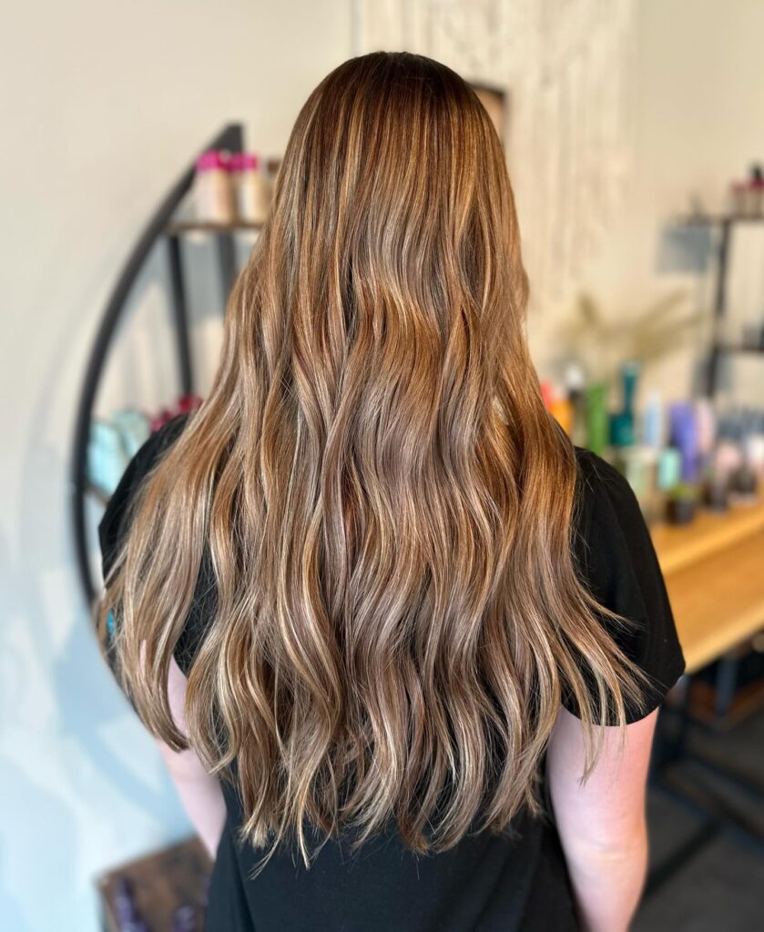 A back view of a woman with long cascading waves in dimensional honey caramel balayage hair.