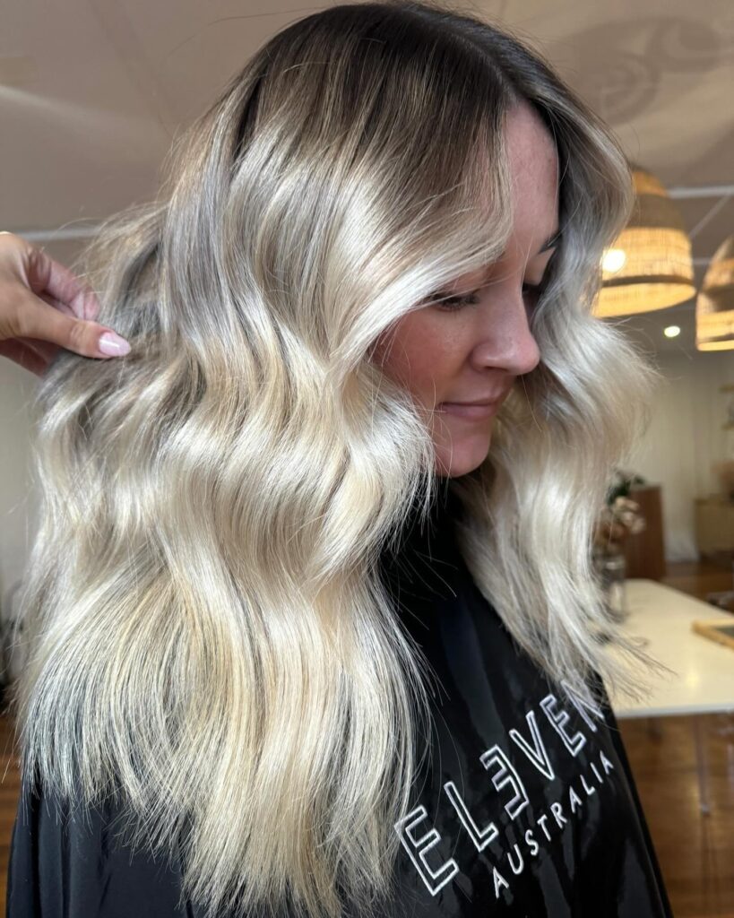 A woman with medium length bright blonde hair featuring a dark shadow root and soft polished waves.