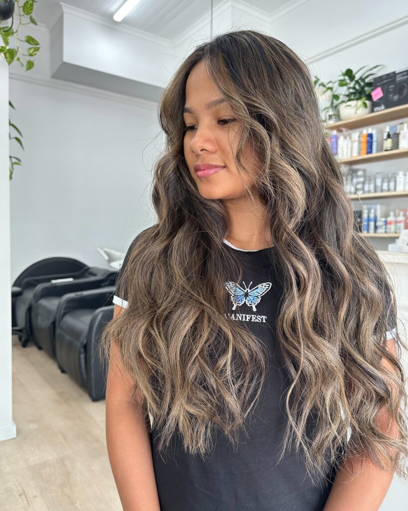 A woman with long wavy dark hair featuring light brown balayage highlights and face-framing layers.