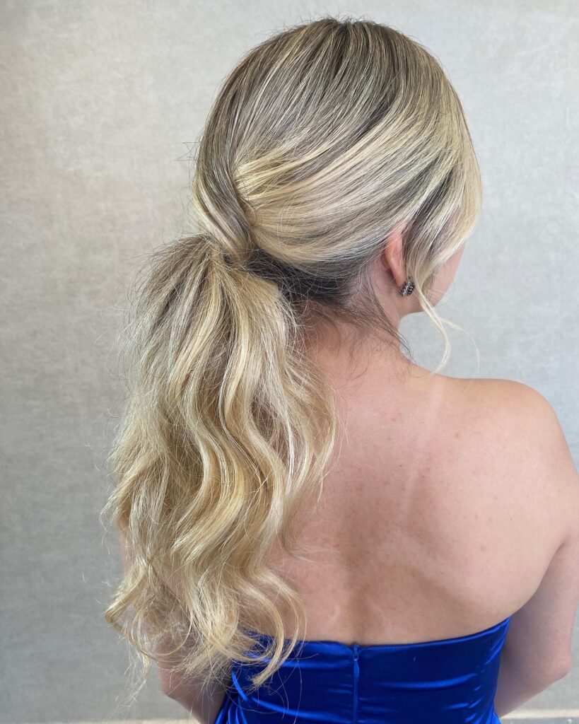 The back view of a woman with highlighted blonde hair styled in a loose low ponytail with a wrap detail and wavy texture.