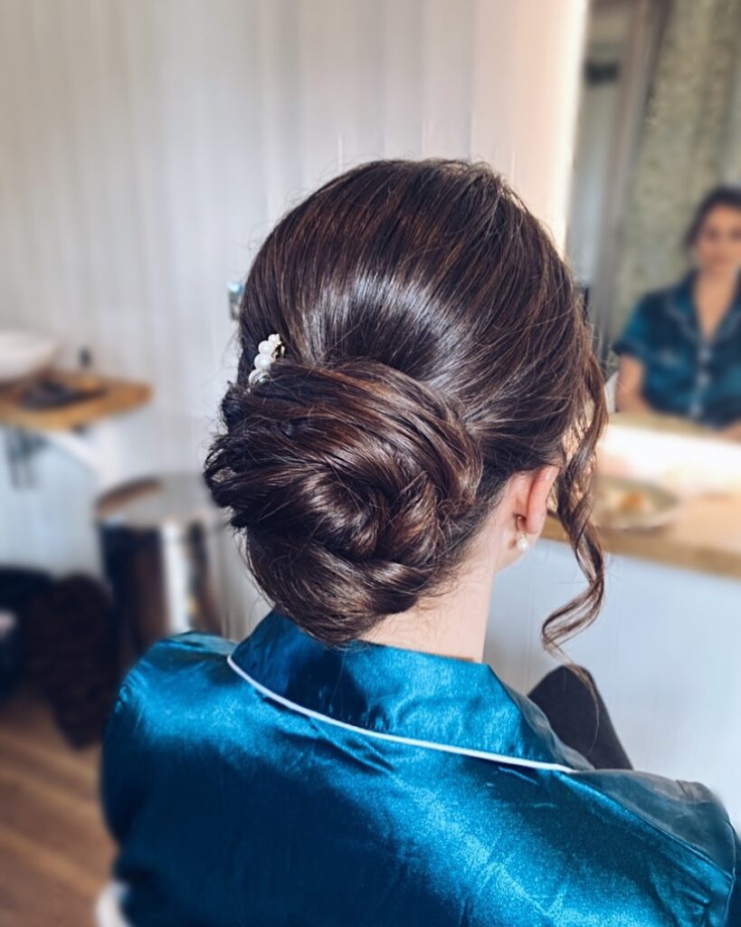 A woman with glossy dark brown hair styled in a structured low chignon decorated with small white pearls and loose side curls.