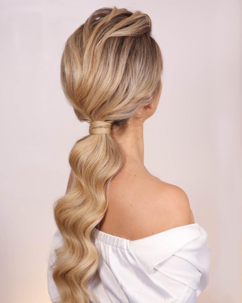 The back view of a woman with long blonde hair styled into a voluminous low ponytail with perfectly defined Hollywood waves and a wrapped base.