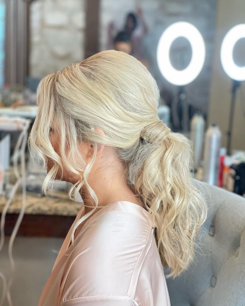 A side view of a woman with platinum blonde hair styled in a wavy low ponytail with curly strands framing her face.