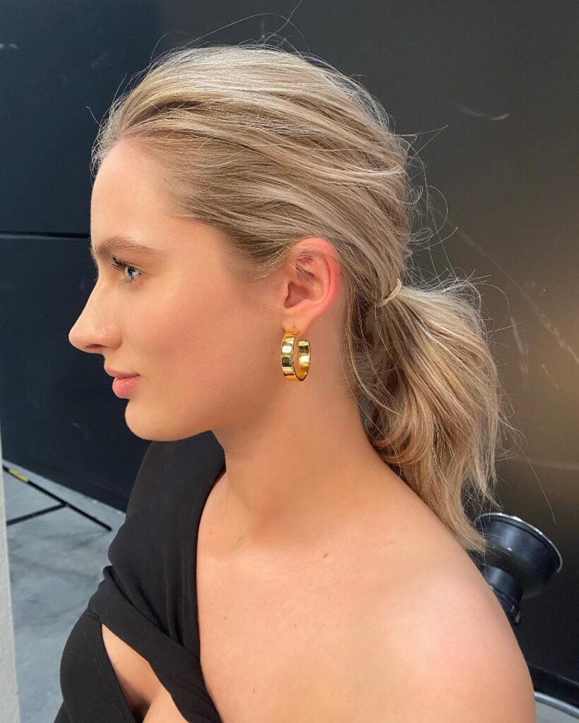 A woman in profile wearing gold hoop earrings and a black top with her blonde hair pulled back into a messy low ponytail.