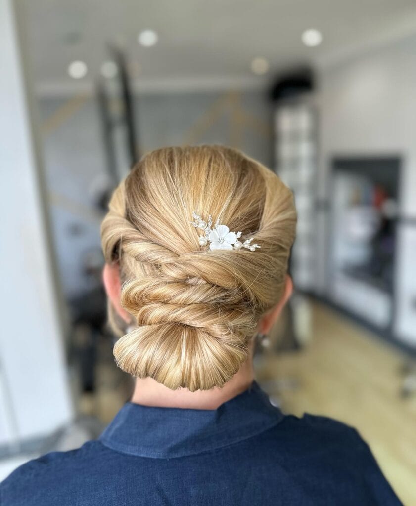 The back of a woman's head showing a golden blonde low bun with twisted sections and a white floral pearl hair clip.