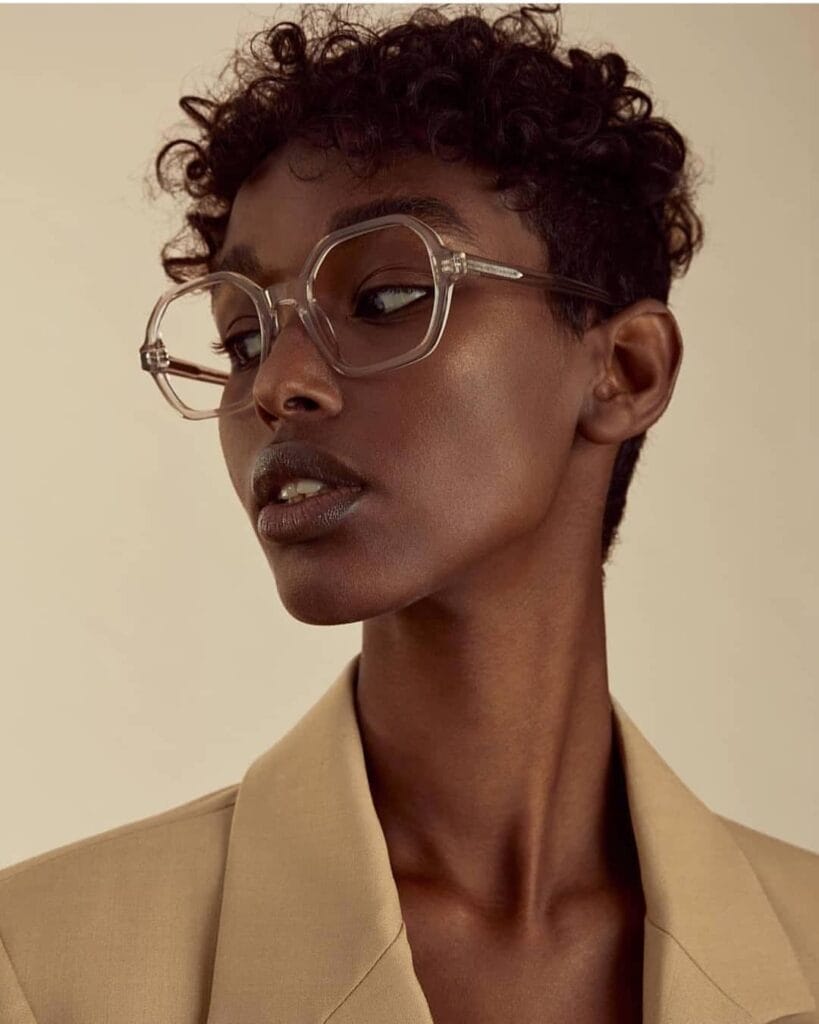 A woman with a short tapered pixie cut featuring tight natural dark curls and wearing stylish clear glasses.