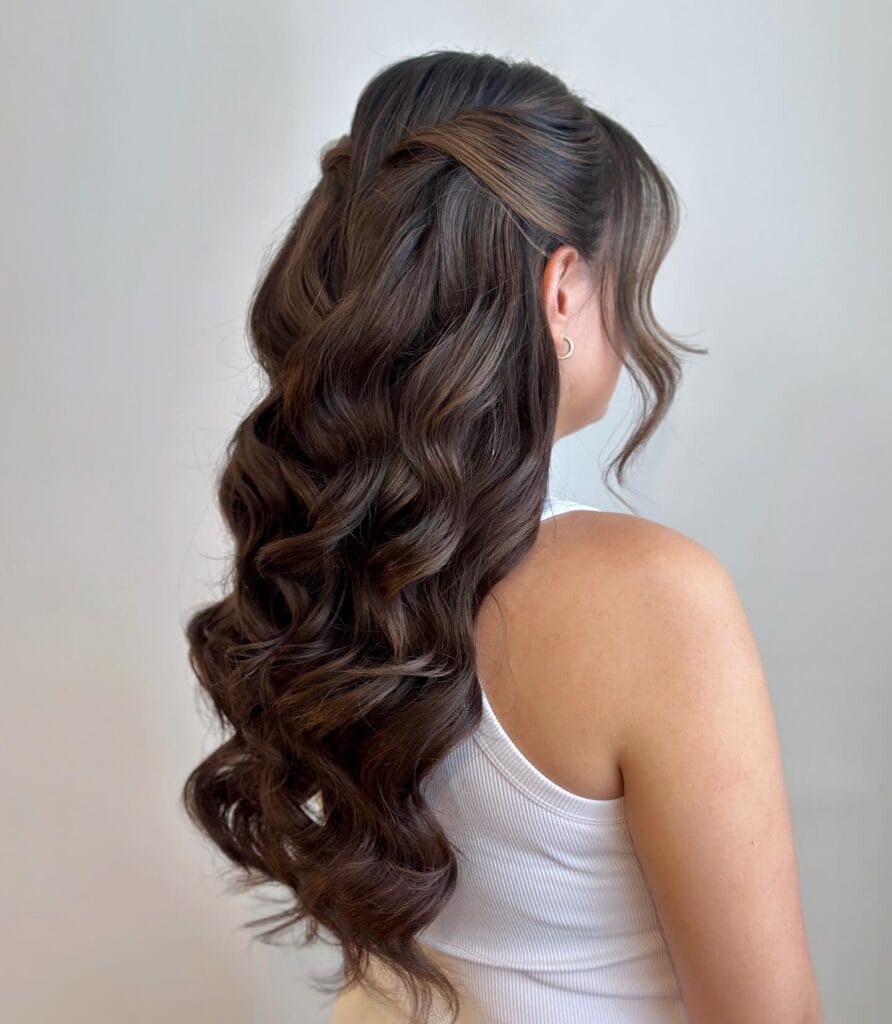 A back view of a woman with long dark brown curly hair in a twisted half up hairstyle with loose front tendrils.