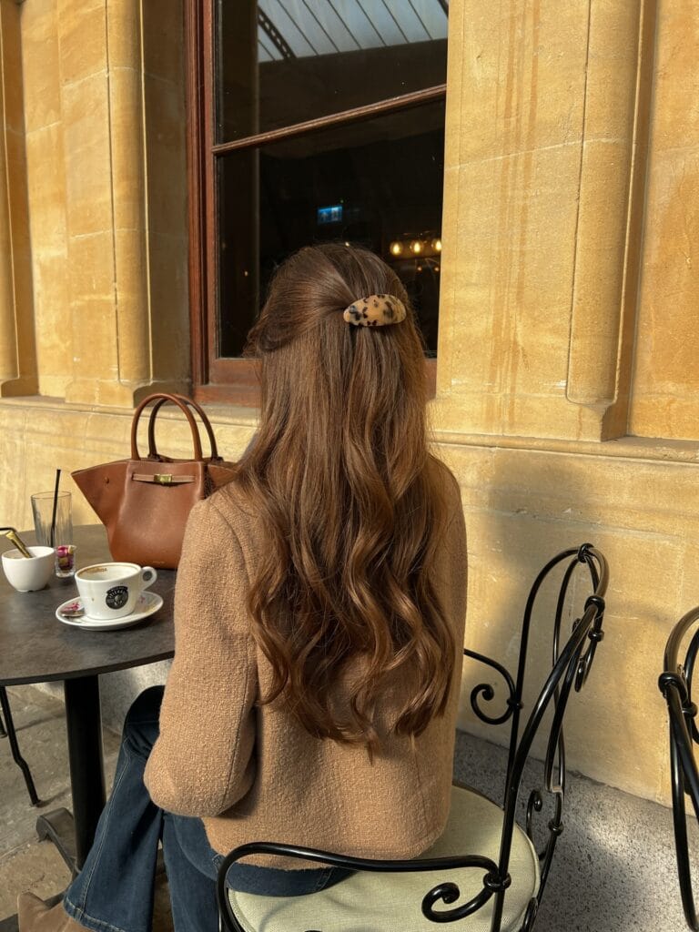A woman sitting at a cafe table from behind showing long wavy brown hair in a half up style secured with a tortoiseshell clip.