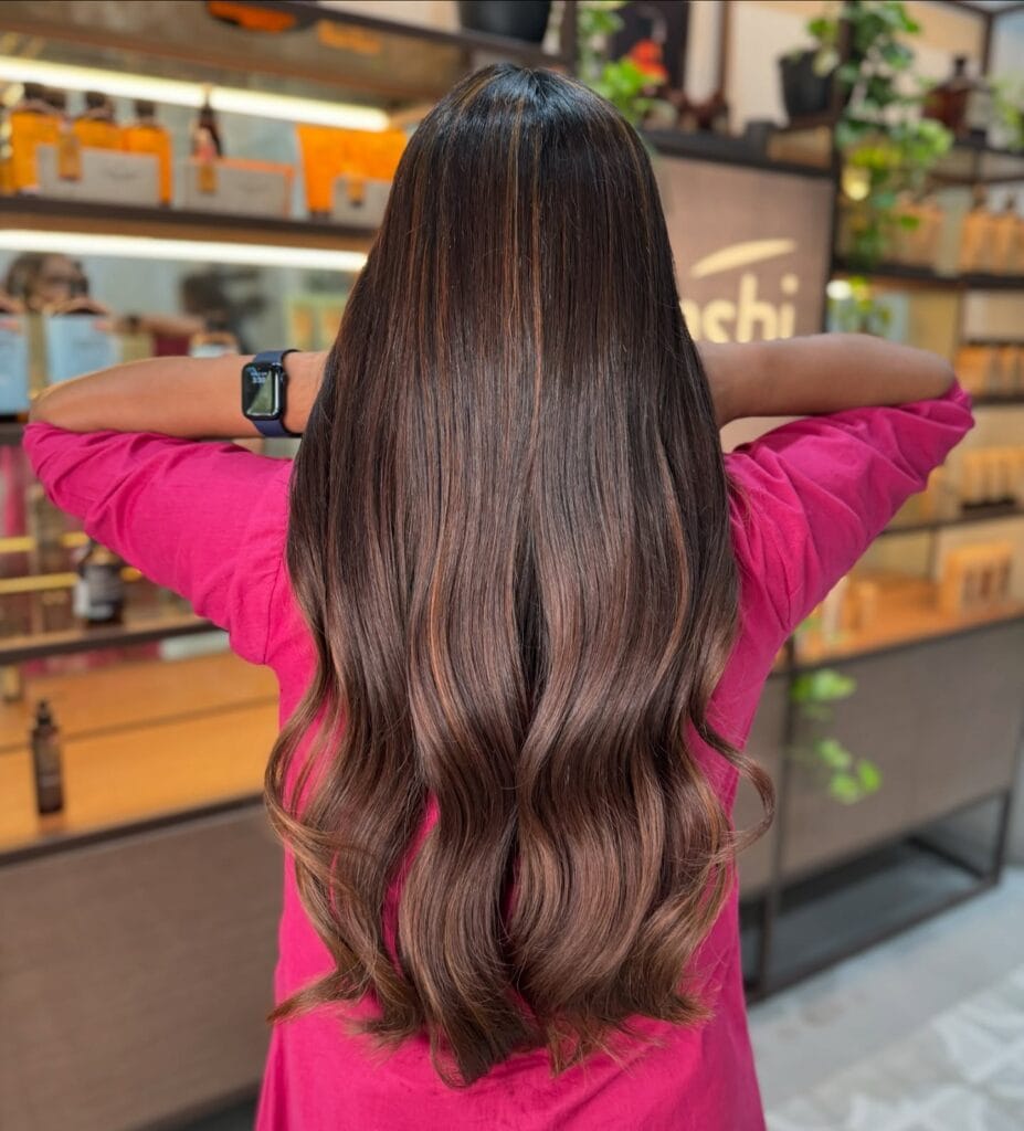 A back view of a woman with long dark brown hair featuring caramel highlights and soft glossy waves.