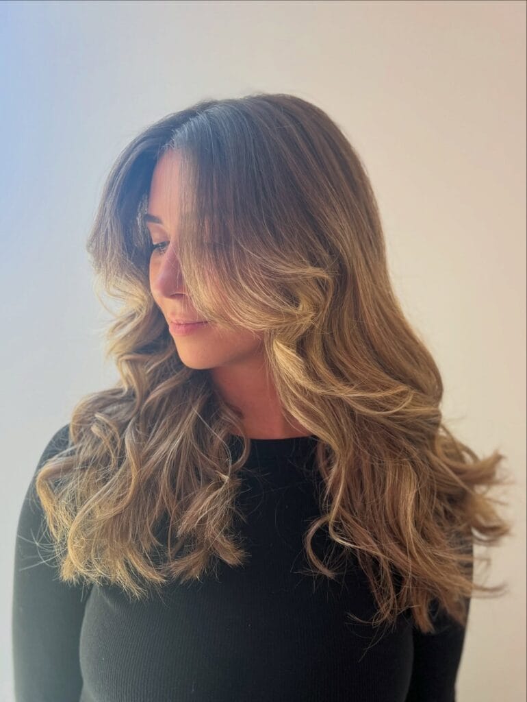 A woman with long layered hair featuring a blonde balayage and voluminous curls that frame her face.