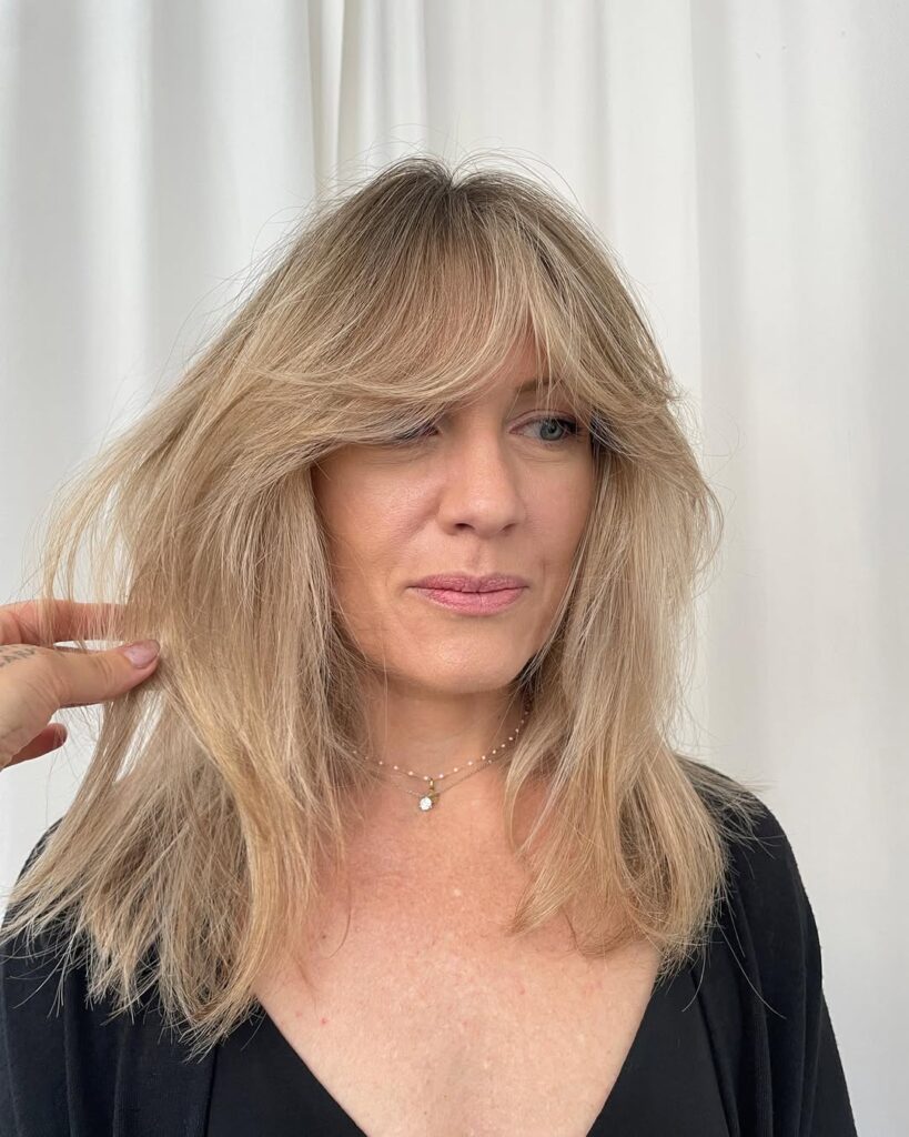 Medium length blonde hair with feathered layers and voluminous curtain bangs framing the face.