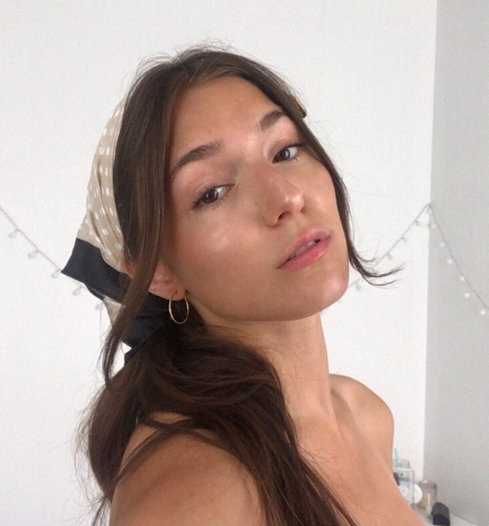 A woman with long wavy brunette hair wears a cream and black polka dot bandana tied as a headscarf with face framing strands.