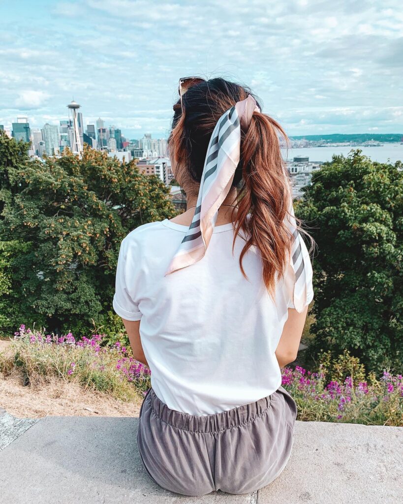 A woman with long wavy brown hair has a high ponytail secured with a satin scarf that has pink, white, and grey stripes.