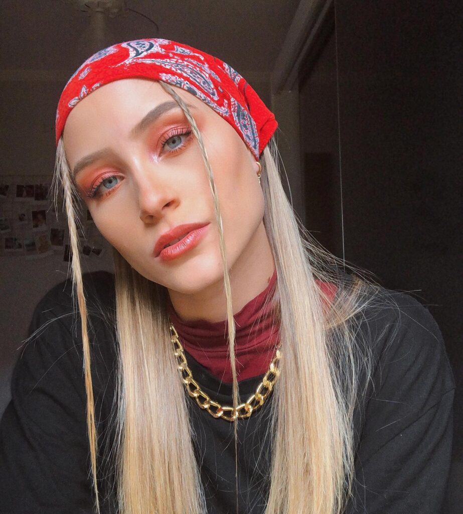 A woman with long, straight platinum blonde hair has a red paisley bandana tied like a headwrap, with two small braids framing her face.
