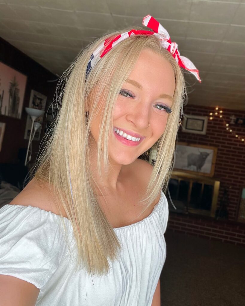 A smiling woman with straight blonde hair has a red, white, and blue patterned bandana tied like a headband around her head, with small tendrils framing her face.