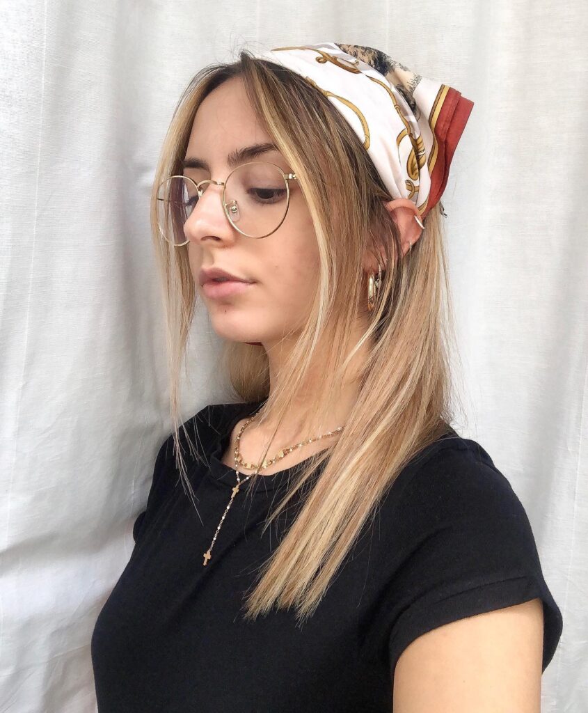 A woman with long highlighted blonde hair and gold glasses wears a silky white and gold patterned bandana tied over her head.
