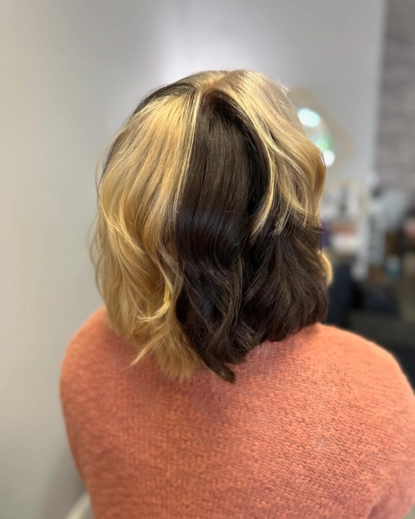 A shoulder-length bob featuring a bold vertical split dye with one half golden blonde and the other half dark brown, styled in soft waves.