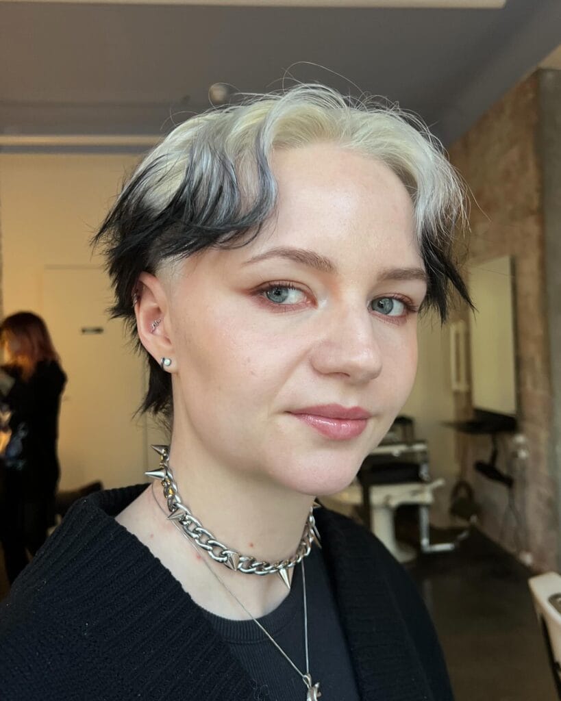 A woman with an edgy two-tone pixie mullet featuring platinum blonde roots and black ends with buzzed sides.