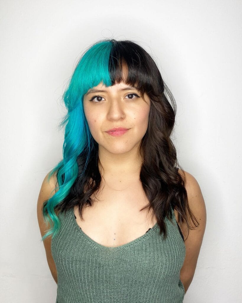 A woman with medium-length wavy hair and full bangs featuring a dramatic vertical split dye with one side vibrant teal and the other side dark brown.