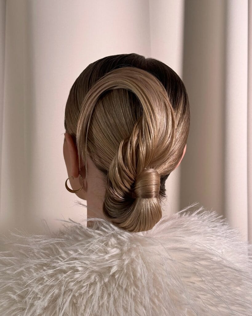 A rear view of a woman with a sleek blonde low chignon featuring a creative and sculptural looped hair design.