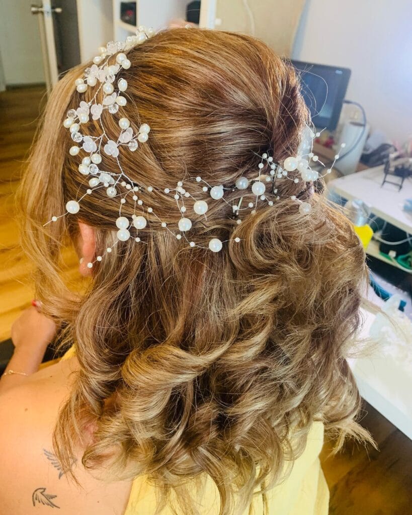 A woman with honey-brown hair in a curly half-updo decorated with a long shimmering pearl and crystal hair vine.
