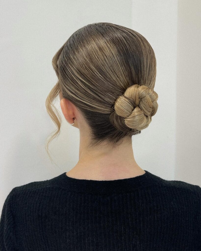 A woman with ash blonde hair styled into a sleek, low braided chignon with loose face-framing waves.
