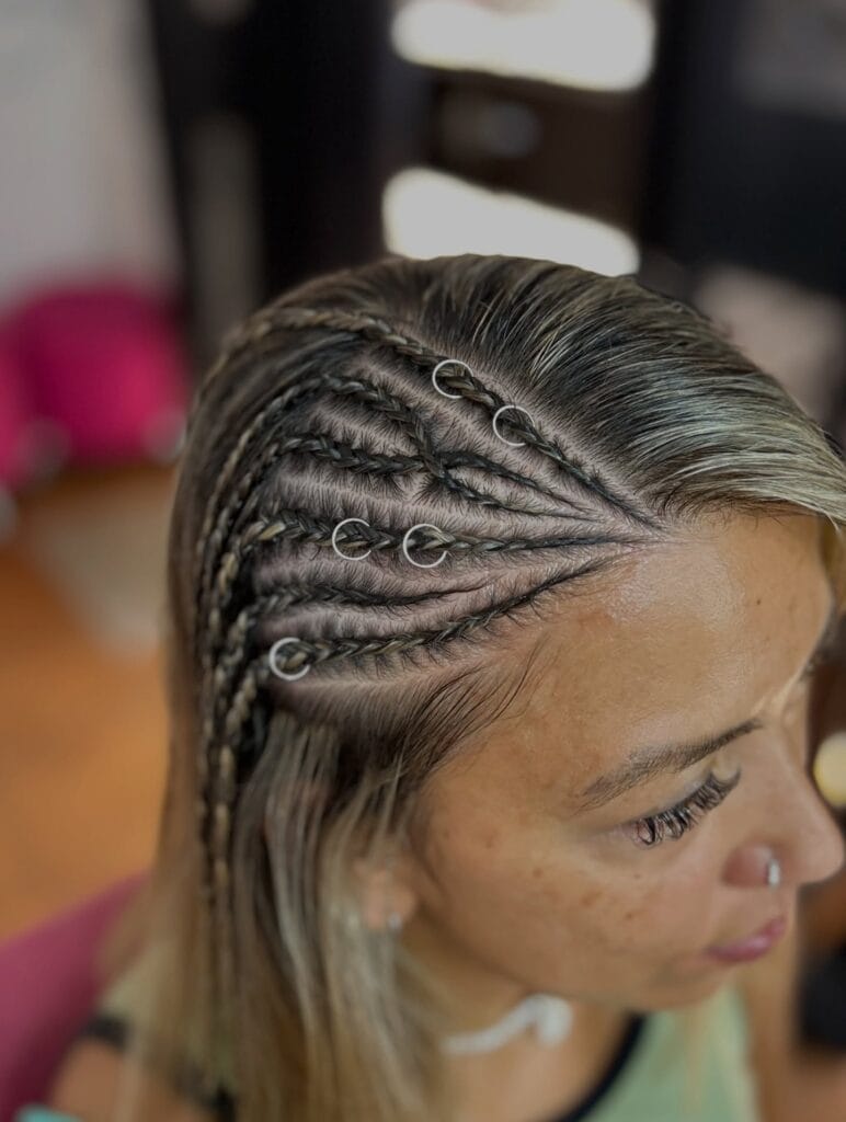 A close-up side view of a woman with several small, tight braids decorated with silver hair rings along the temples.